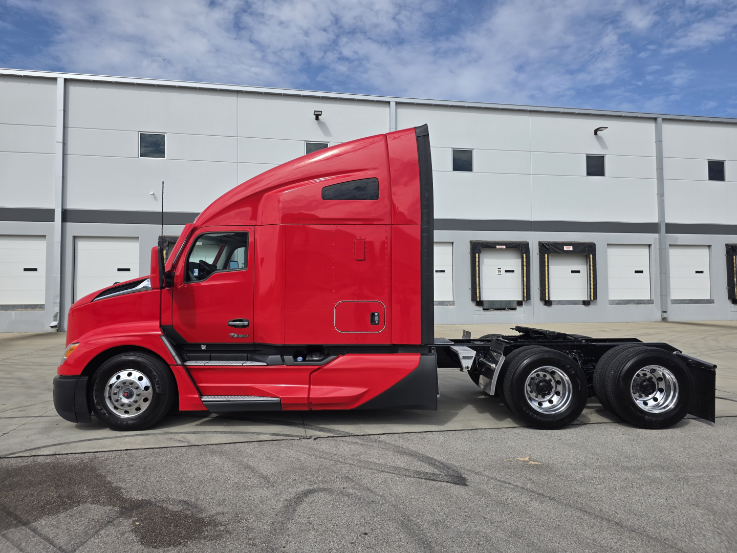 Kenworth T680 tractor in red side view showing sleeper cab, chassis, and rear wheels with fifth wheel; daytime with industrial building in background.
