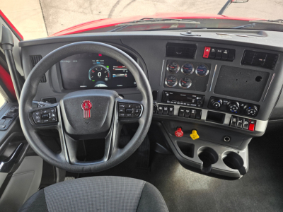 Dashboard view of a 2023 Kenworth T680 cab showing steering wheel with Kenworth emblem, instrument cluster, and control panels; multiple gauges, switches, and cup holders visible.