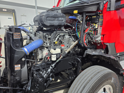 Kenworth T680 engine bay showing 6-cylinder diesel engine with blue intercooler hose, yellow oil cap, wiring harnesses, and front wheel visible.
