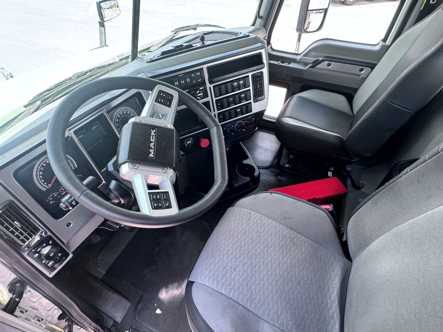 Truck cab interior showing steering wheel with Mack badge and dashboard controls, driver's seat, passenger seat, center console, and windshield view.