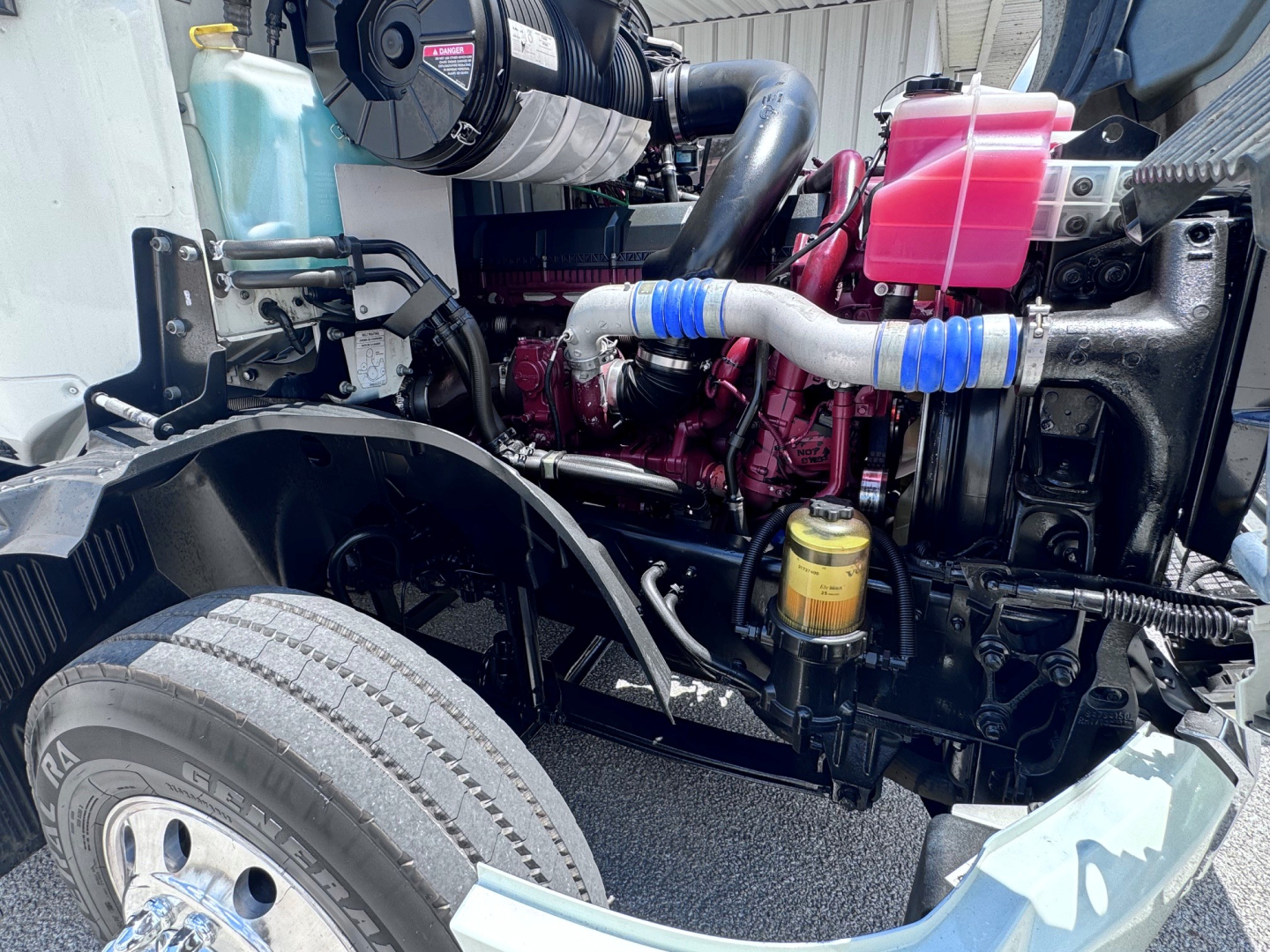 Engine bay of a 2020 Mack Anthem showing coolant reservoir, air intake, pink coolant reservoir, blue silicone hoses, yellow oil filter, and exposed engine components.