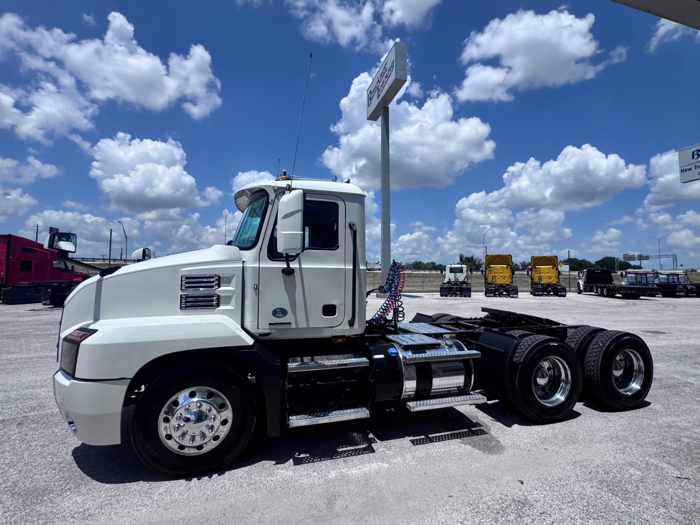 2020 Mack Anthem day cab tractor in a parking lot with clear blue sky. Side view shows white cab, dual rear axles, chrome fuel tanks, and exhaust. No visible text on cab apart from reflections.