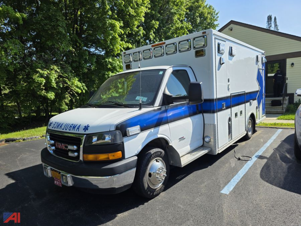 GMC Savana ambulance front-left view with blue and white livery, multiple rooftop medical compartments, and ‘AMBULANCE’ text on the cab grille area.