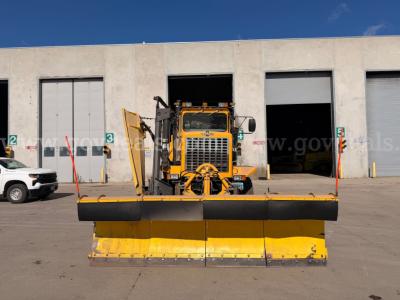 Front view of a yellow Oshkosh snow plow blade attached to a front-end loader in a warehouse yard.