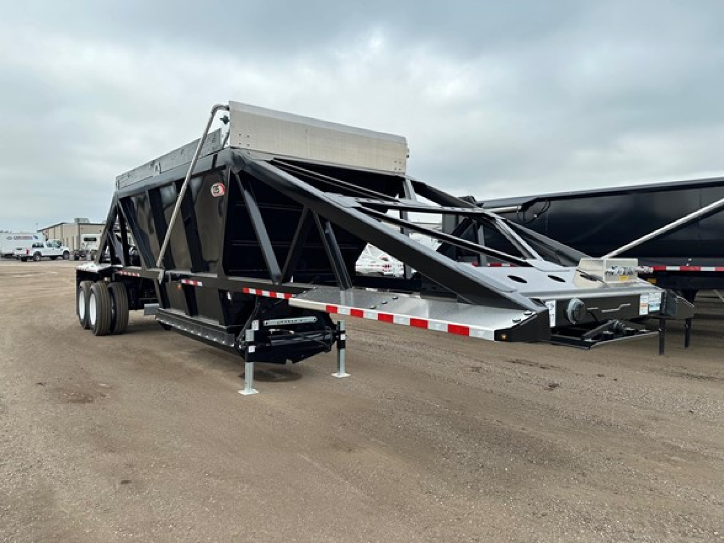 Bottom-dump trailer on a dirt lot, black frame with triangular truss sides, extended front hopper, tandem rear wheels, landing gear and support legs visible.