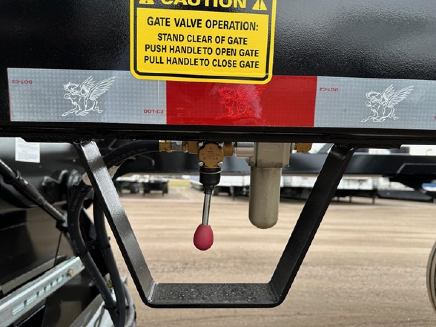 Bottom dump trailer hitch area with yellow caution sticker and red/white reflective tape, shows valve handle and pink knob, part of frame undercarriage.