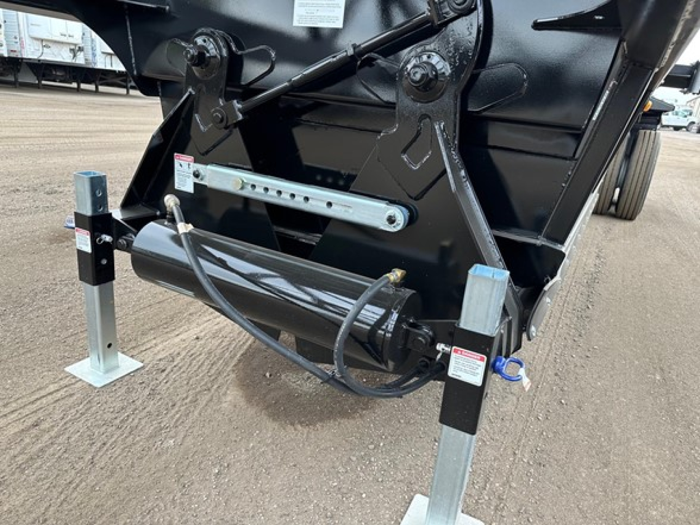 Bottom-dump trailer hitch area showing hydraulic cylinder, support legs, and frame with pins and hydraulic hoses.