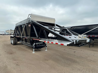 Bottom-dump trailer on a dirt lot, black frame with triangular truss sides, extended front hopper, tandem rear wheels, landing gear and support legs visible.