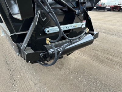 Trailer bottom dump hitch area with hydraulic hoses and metal bracket; visible text on a small label and bolts, part of the dump trailer frame.