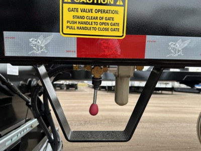 Bottom dump trailer hitch area with yellow caution sticker and red/white reflective tape, shows valve handle and pink knob, part of frame undercarriage.