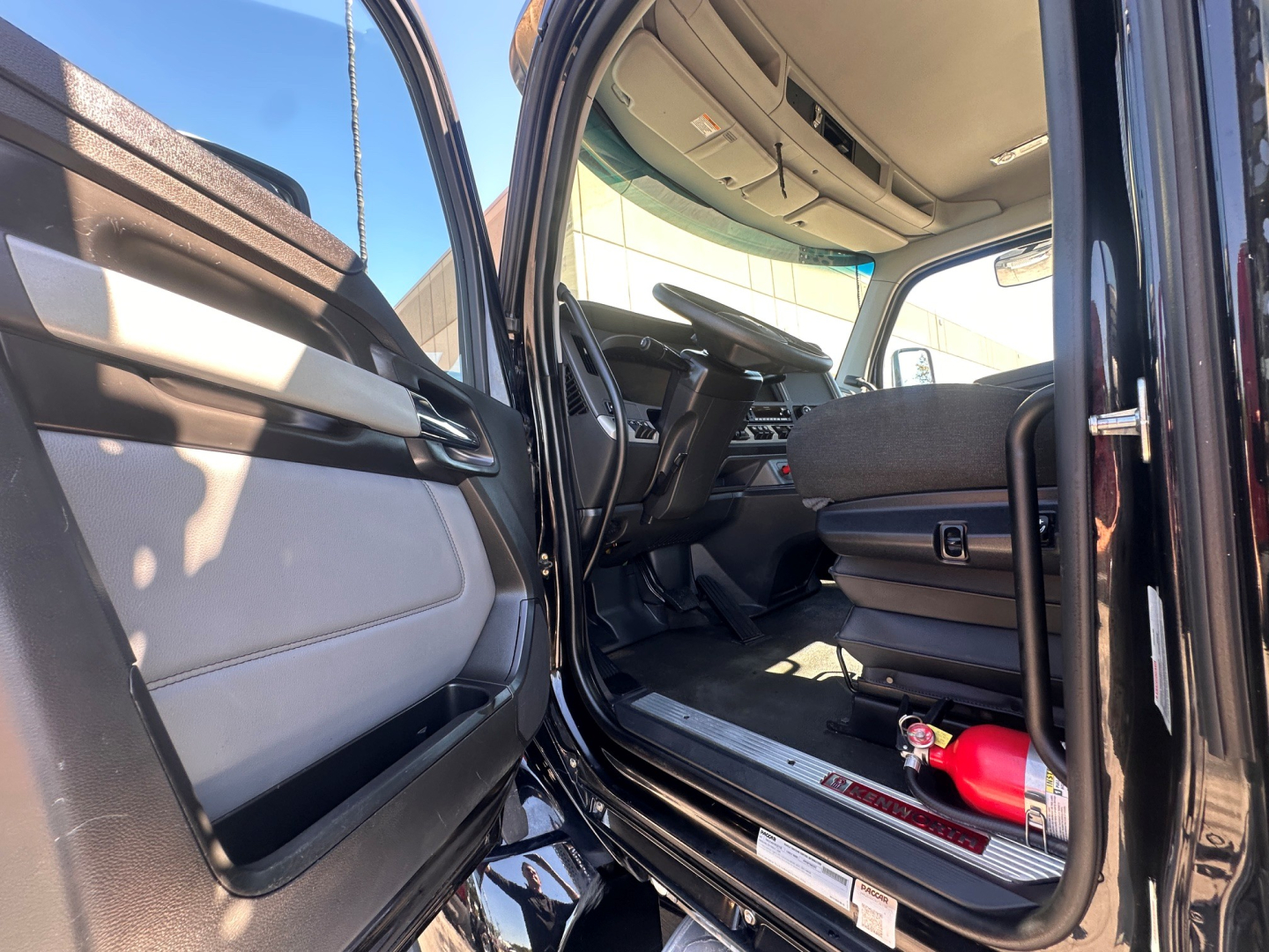 Kenworth T880 truck cab interior with open driver door showing seats and dashboard; floor entry with ladder steps and a small red fire extinguisher near the seat.