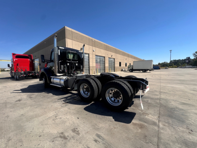 Kenworth T880 tractor in a parking lot, side view showing dual rear wheels and fifth-wheel, with a beige warehouse in background.