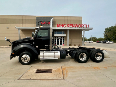 Kenworth T880 truck in profile view with three-axle rear, black cab, chrome exhaust stack, fuel tanks, and MHC Kenworth dealership building in background.