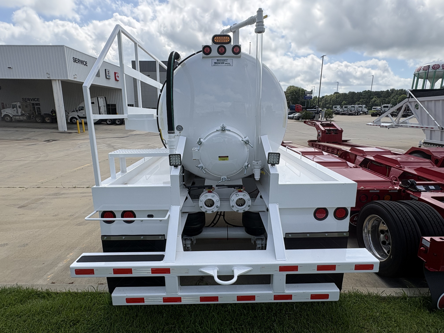 Rear view of a white vacuum trailer with a circular tank, rear lights, and hoses. Text and labels on the tank include warning decals and fittings. Industrial lot in background.