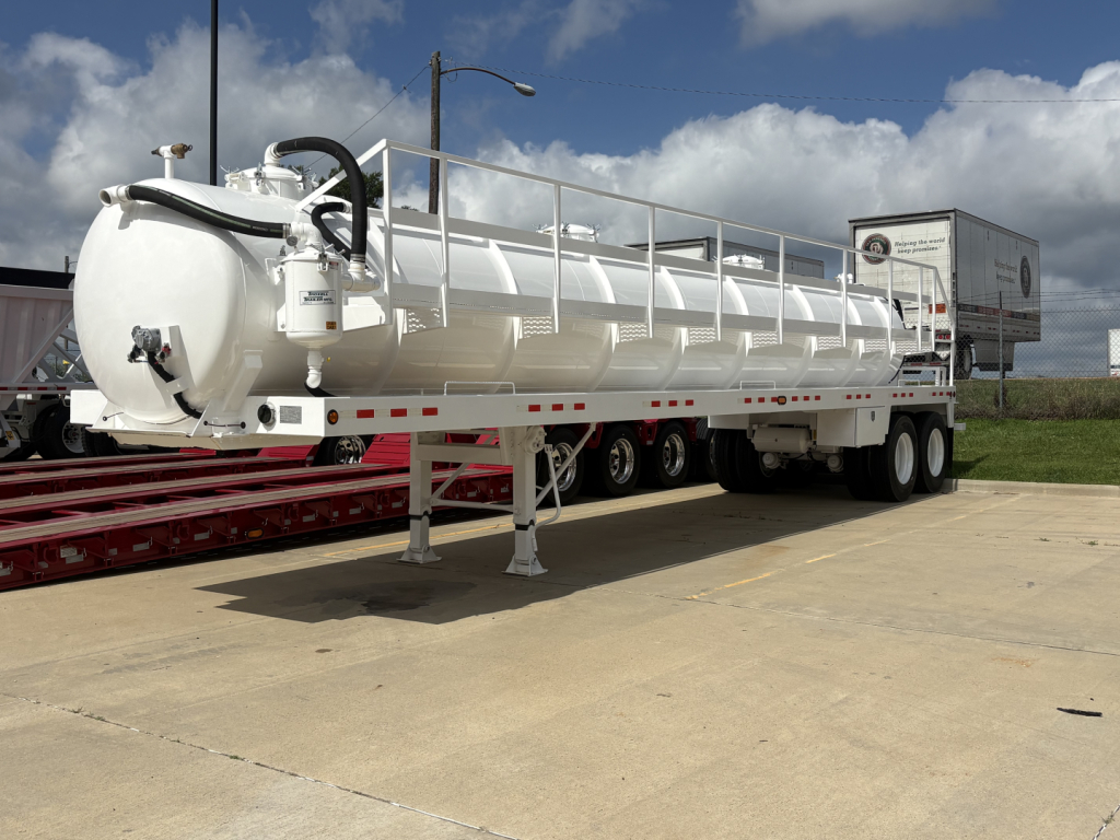 Large white vacuum trailer with cylindrical tank and rear wheels, hoses and access ladder on top, parked in lot.