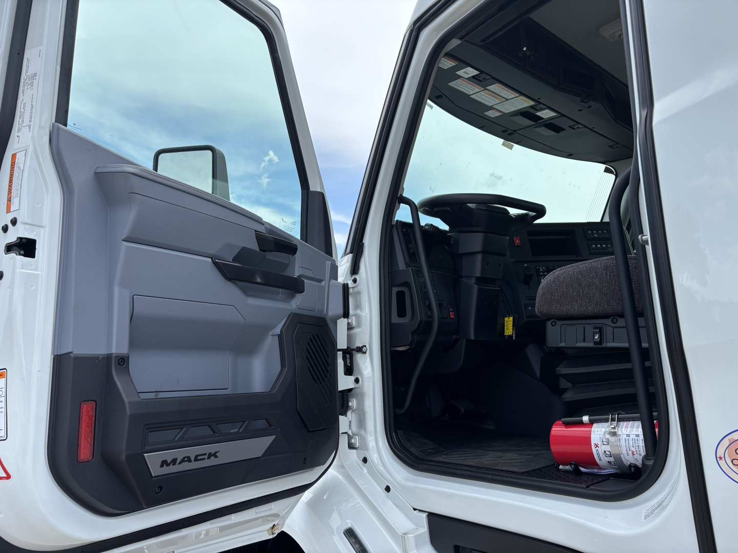 Truck cab interior showing steering wheel, dashboard, and open door with Mack logo on door panel; red extinguisher on floor.