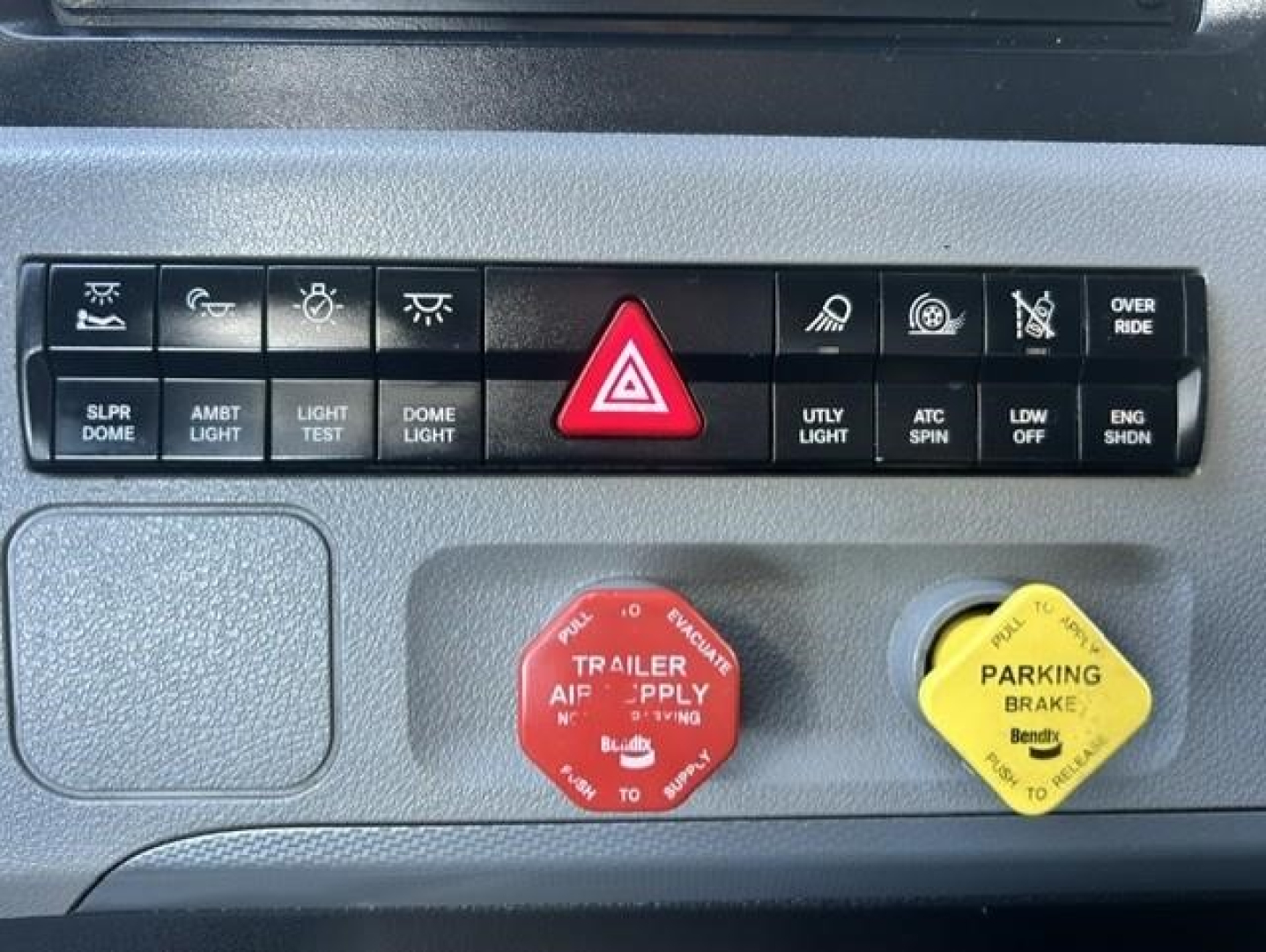 Freightliner Cascadia 126 dash area showing control switches, red hazard triangle button, red trailer air supply knob, and yellow parking brake knob.