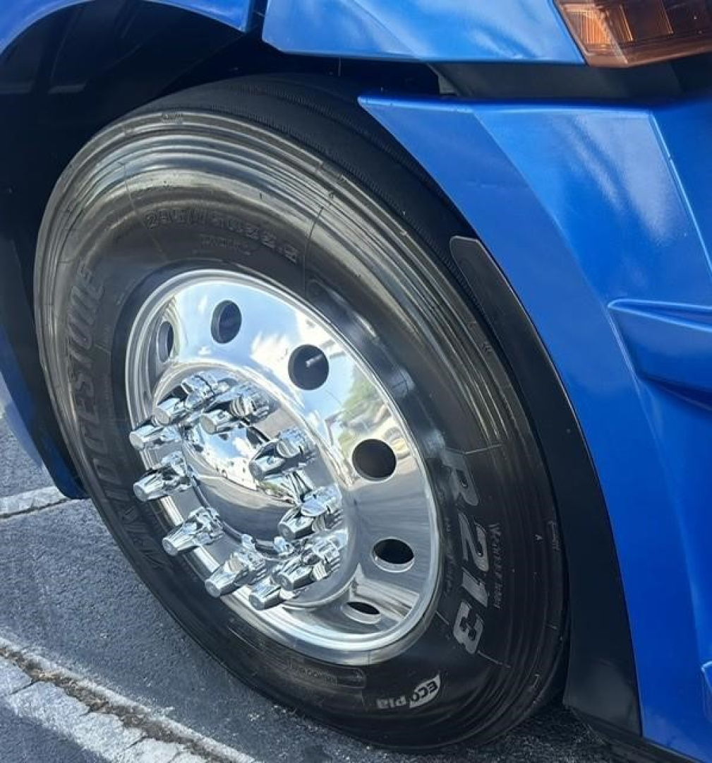 Truck front wheel well showing a polished chrome wheel with multiple lug nuts on a blue Freightliner Cascadia 126; tire sidewall shows tire model and branding.