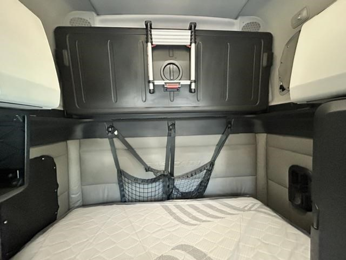 Freightliner Cascadia 126 sleeper cab interior cargo area with storage cabinet overhead and bed in lower bunk, cords and net storage visible.