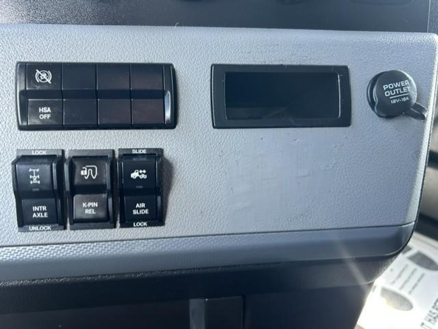 Dashboard panel of 2022 Freightliner Cascadia showing multiple switches and power outlet with round cap on the right.