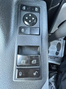 Control panel with window/mirror adjustment pad and lock/unlock switches inside a Freightliner Cascadia cab. Visible buttons include a circular directional pad with four arrows, two small rectangular buttons above, two window control switch