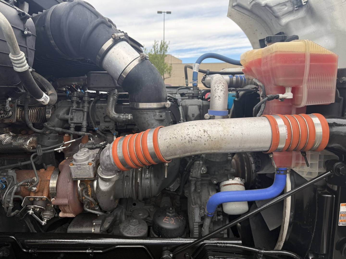 Engine bay view of a 2020 Kenworth T680 conventional daycab showing turbocharger, intercooler piping, coolant reservoir, and various hoses.