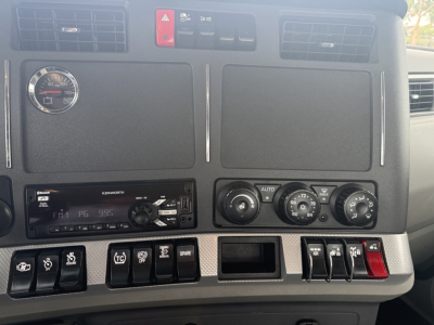 Dashboard of a Kenworth T680 showing a round gauge, two large vent panels, controls, and a Kenworth radio/display; multiple switches and climate controls visible.