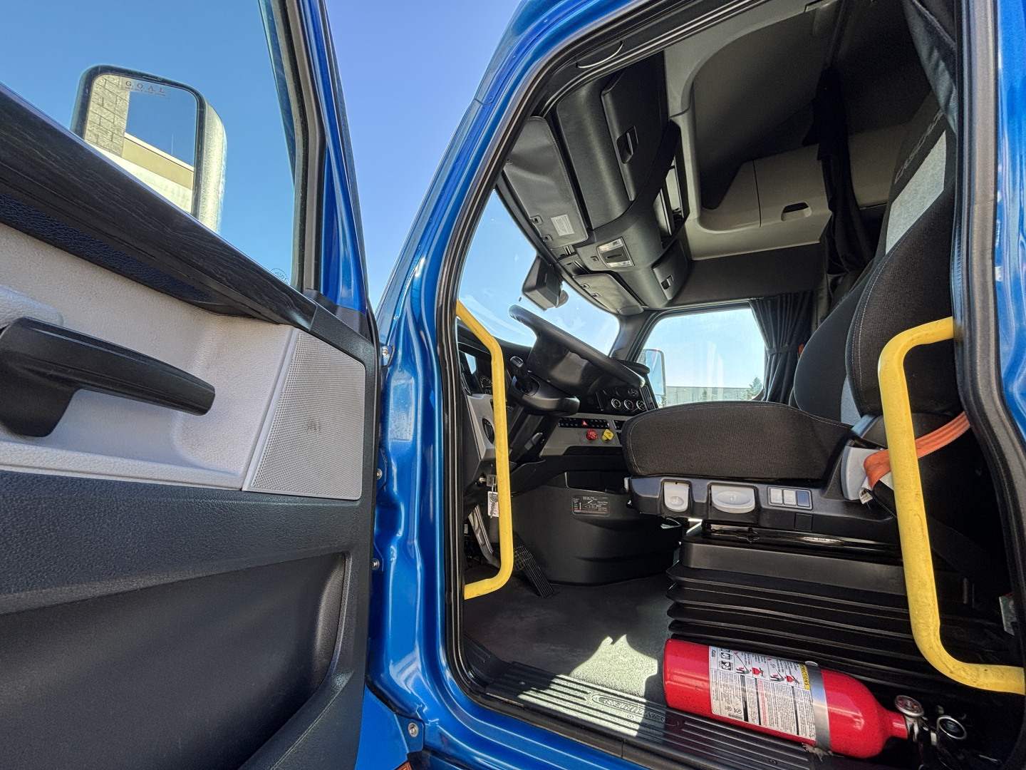 Truck cab interior view from open driver door showing steering wheel, dashboard, and seats; safety bar and fire extinguisher mounted near floor.