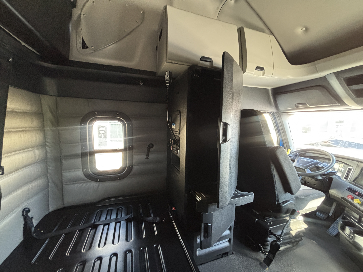 Cab interior of a Freightliner PT126064ST showing driver seat, steering wheel, center console, and sleeper area with window and storage cabinet.