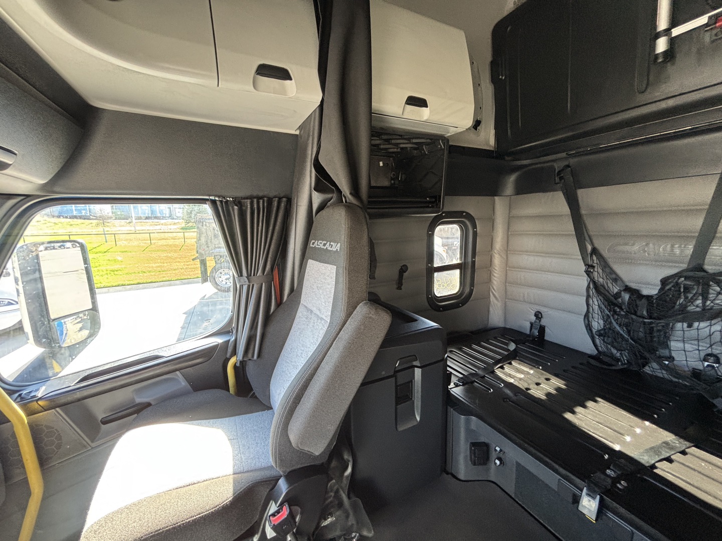 Cab interior of a Freightliner truck sleeper with driver seat, overhead cabinets, and bunk area visible; windows show outside field.