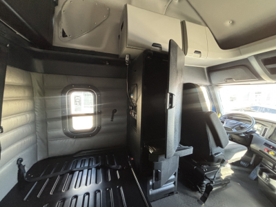 Cab interior of a Freightliner PT126064ST showing driver seat, steering wheel, center console, and sleeper area with window and storage cabinet.