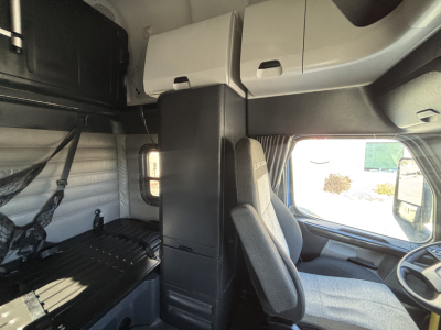 Truck cab interior showing driver's seat and sleeper area with overhead storage cabinets and a window. Text not clearly legible.