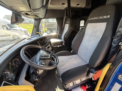 Truck interior with steering wheel, dashboard, and two Gravely seats labeled Cascadia; view toward cab alley and sleeper area; dashboard and control panel visible.