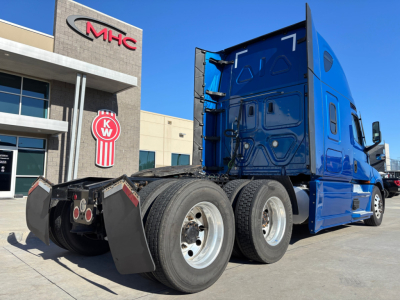Truck rear and side view showing tandem axle, blue sleeper cab, and fifth-wheel area. Visible tires, chassis, and mud flaps with a dealership building in background.