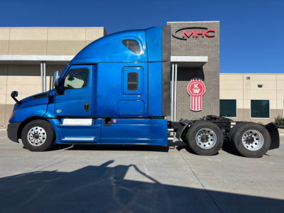 Truck cab and chassis along side view showing blue Freightliner PT126064ST with sleeper cab and KW sign in background.