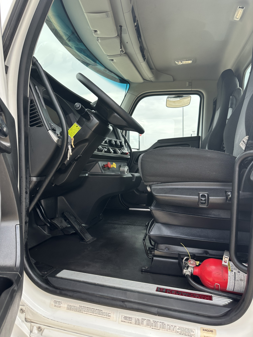 Truck cab interior view of a 2022 Kenworth T880 daycab showing driver seat, dashboard, steering wheel, pedals, and a fire extinguisher near the door.