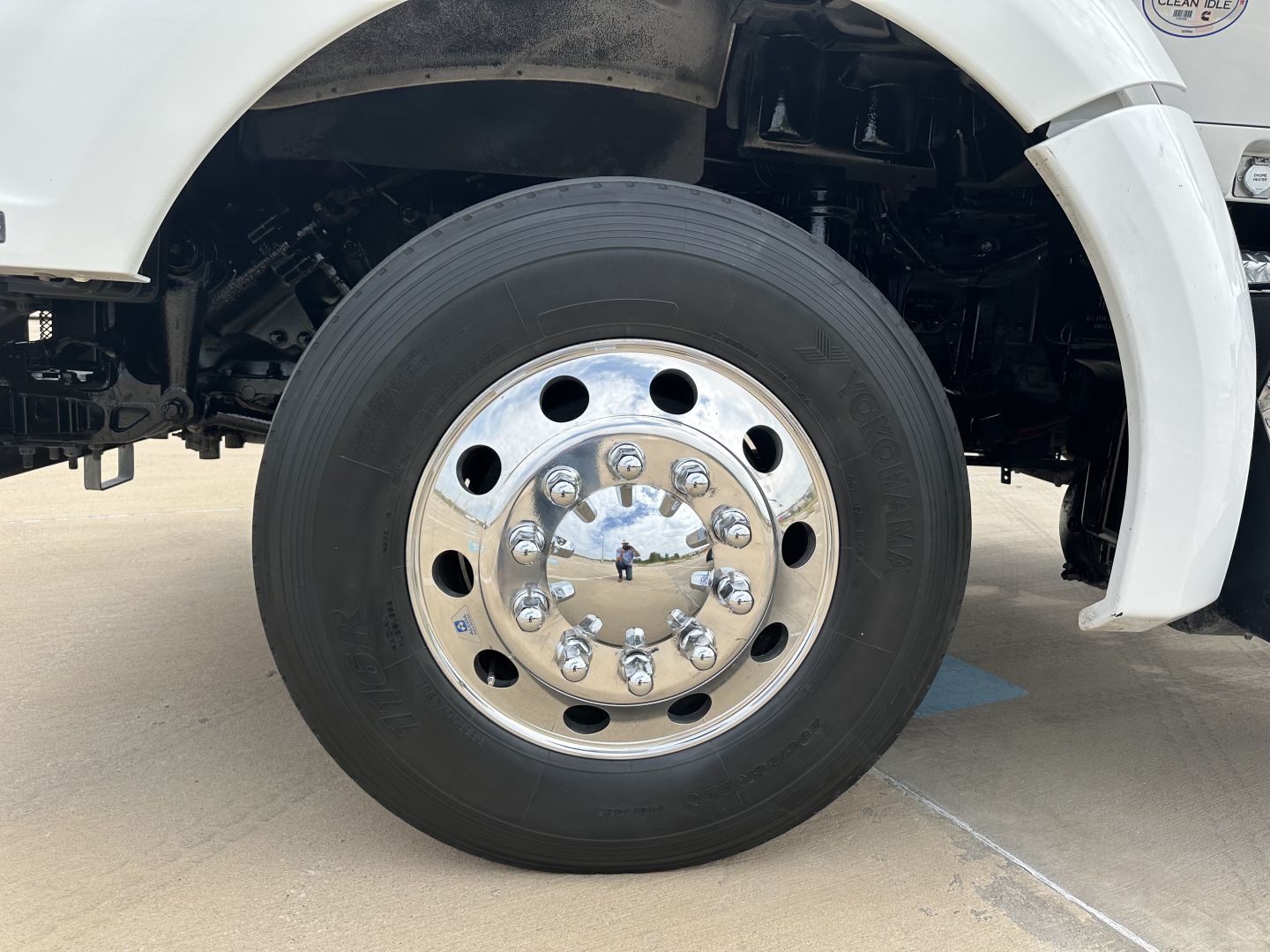 Kenworth T880 wheel and front axle area showing steel hub with lug nuts and tire, portion of white cab/body in background.