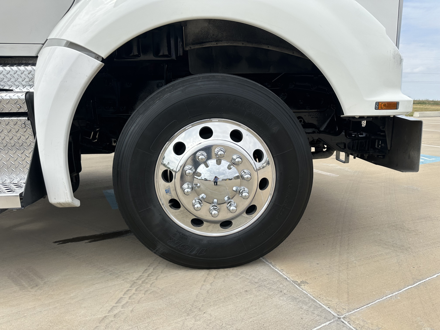 Front wheel of a Kenworth T880 truck showing a polished metal hub and black tire; part of white fender and step visible.