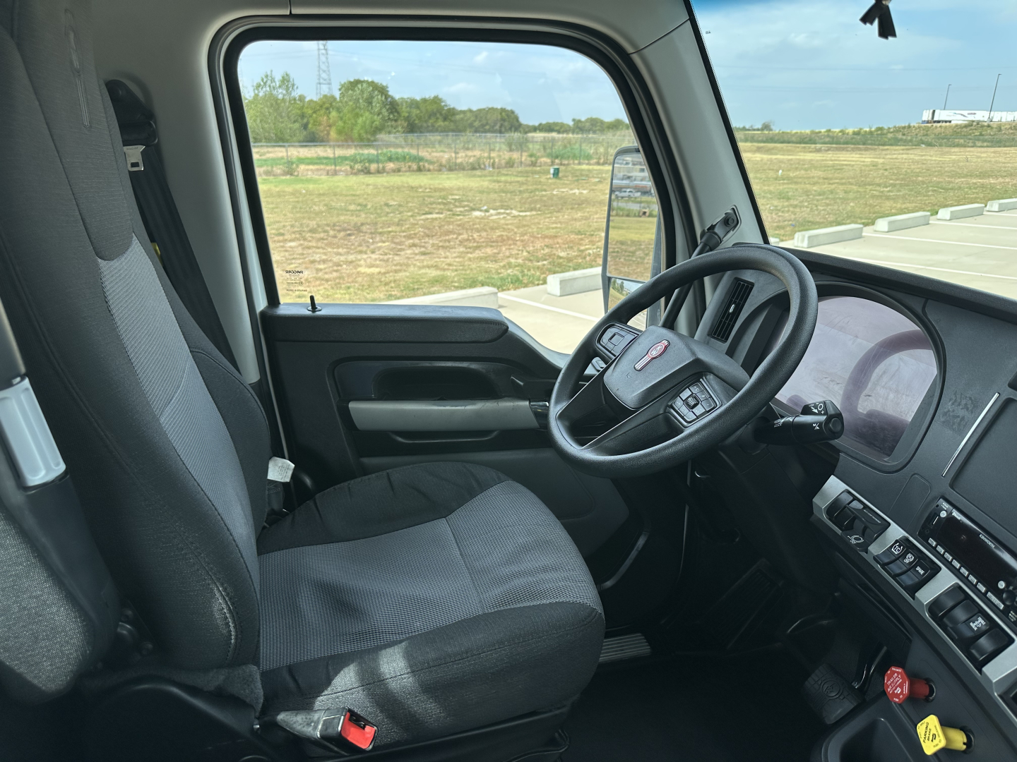 Truck cab interior showing steering wheel with Kenworth logo, driver seat, and dashboard; window view of open field.