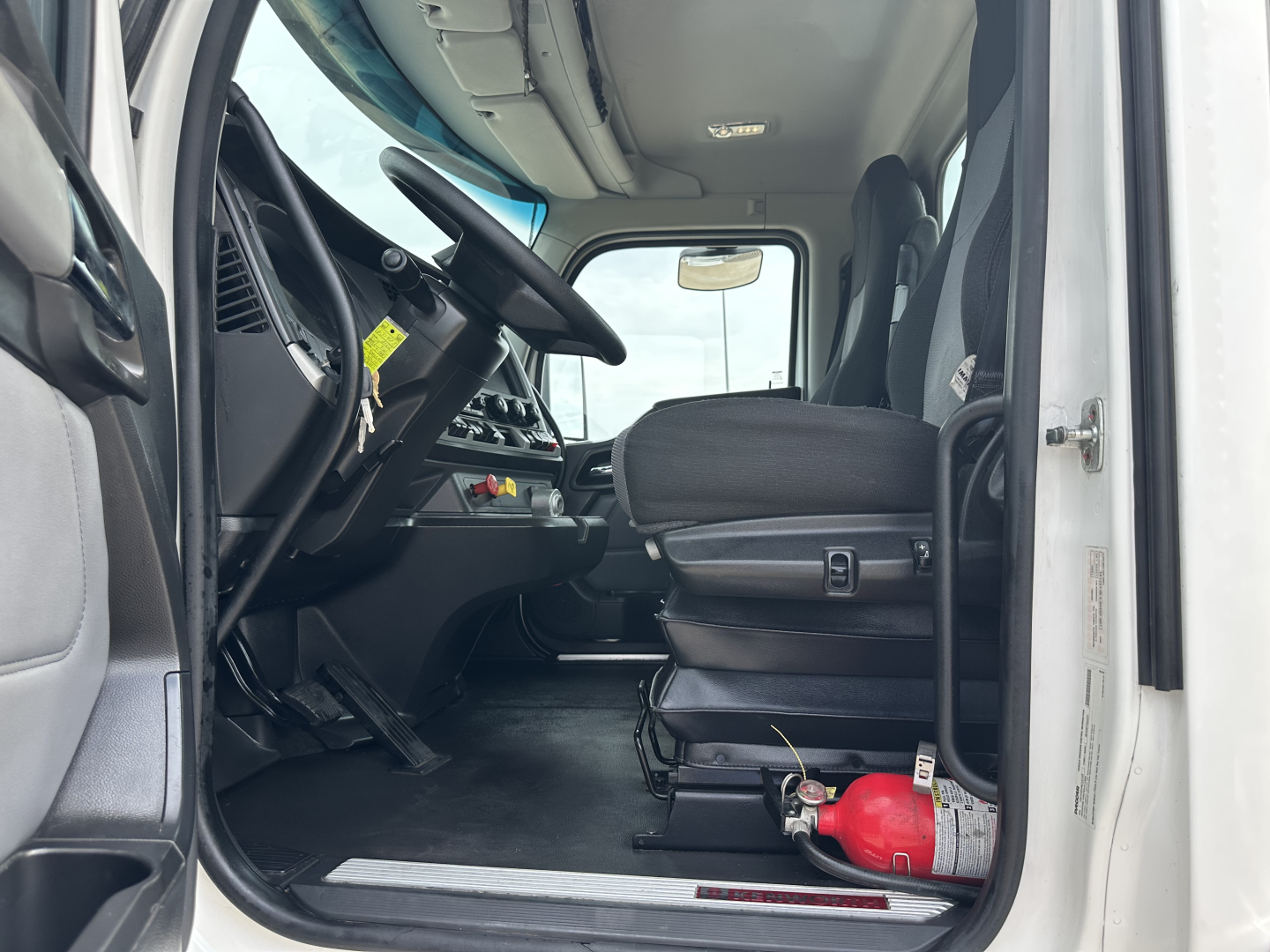 Kenworth truck interior with driver seat visible, steering wheel, dashboard controls, and a red fire extinguisher near the floor. Side door open.