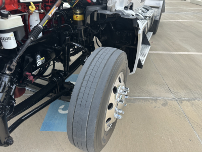Kenworth T880 front wheel and engine bay area showing drivetrain components and suspension; near-side front wheel with exposed frame and steps; visible hydraulic lines and reservoir area.