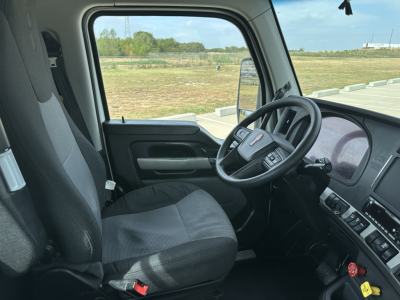 Truck cab interior showing steering wheel with Kenworth logo, driver seat, and dashboard; window view of open field.