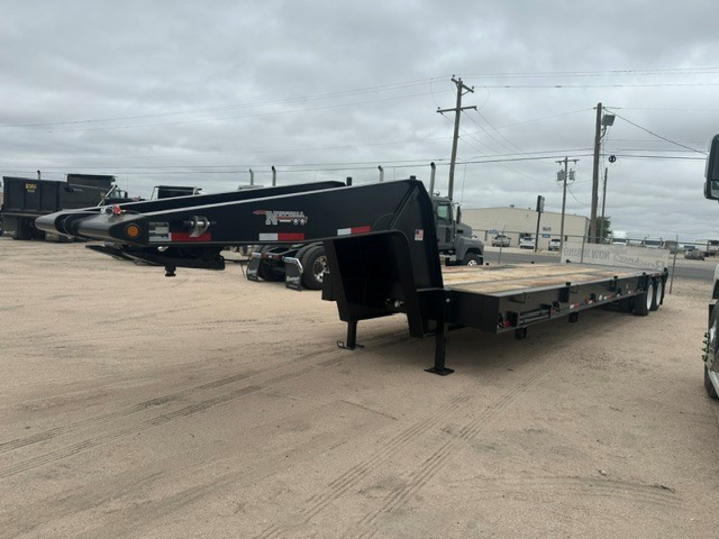 New lowboy trailer with black frame and long flatbed, front gooseneck area, landing legs visible, parked on dirt lot.