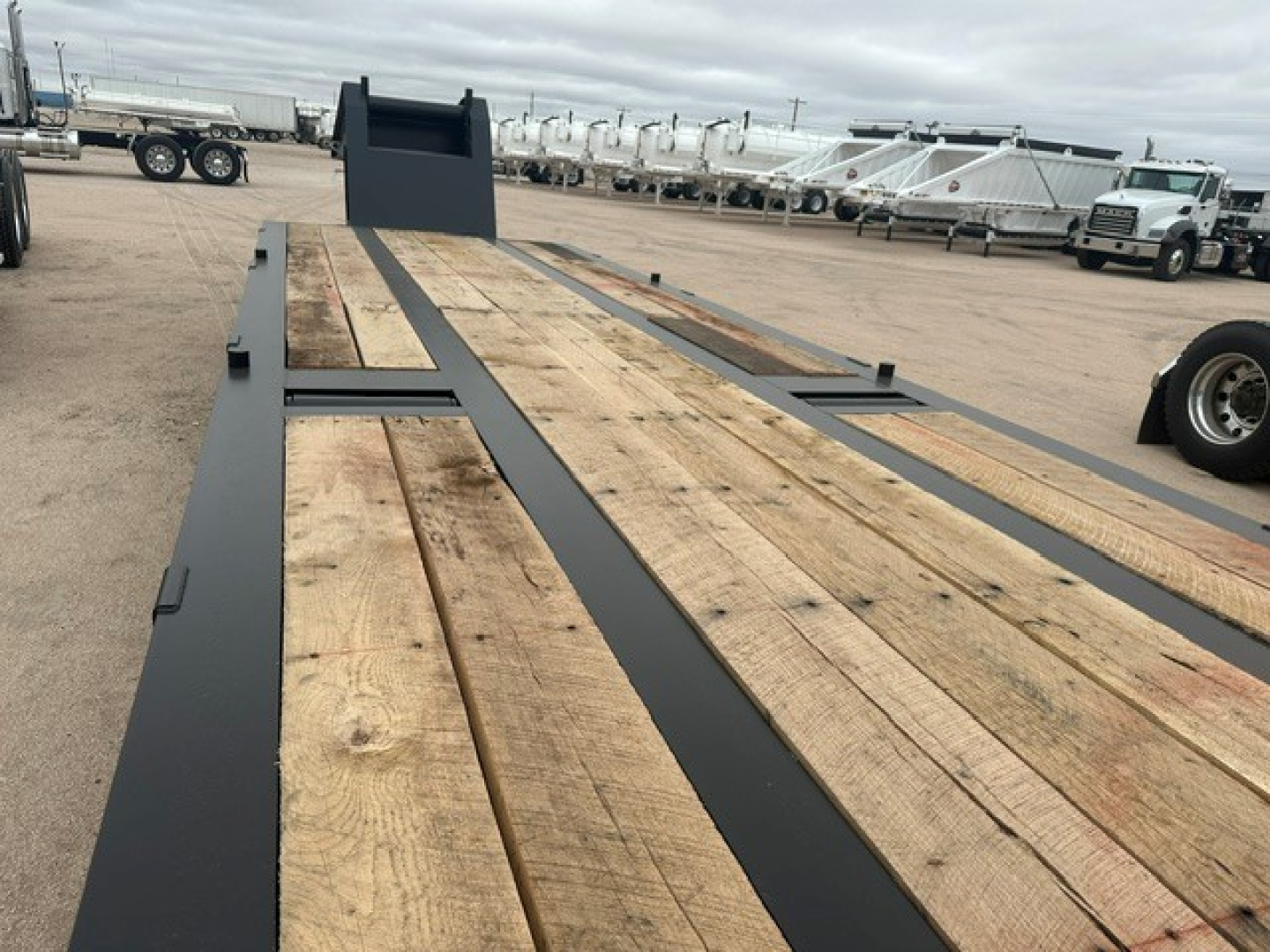 Trailer flatbed showing wooden deck boards with black steel frame and ramps in background; long lowboy trailer in an open lot.