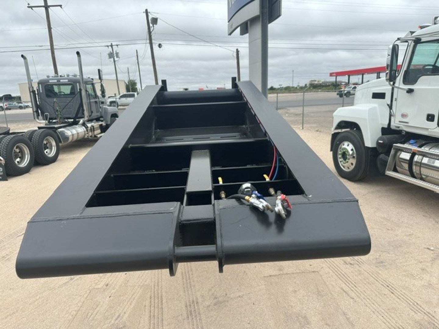 Lowboy trailer bed showing open frame with hydraulic lines and coupler fittings, black paint, steel structure, flat deck area, industrial yard in background.