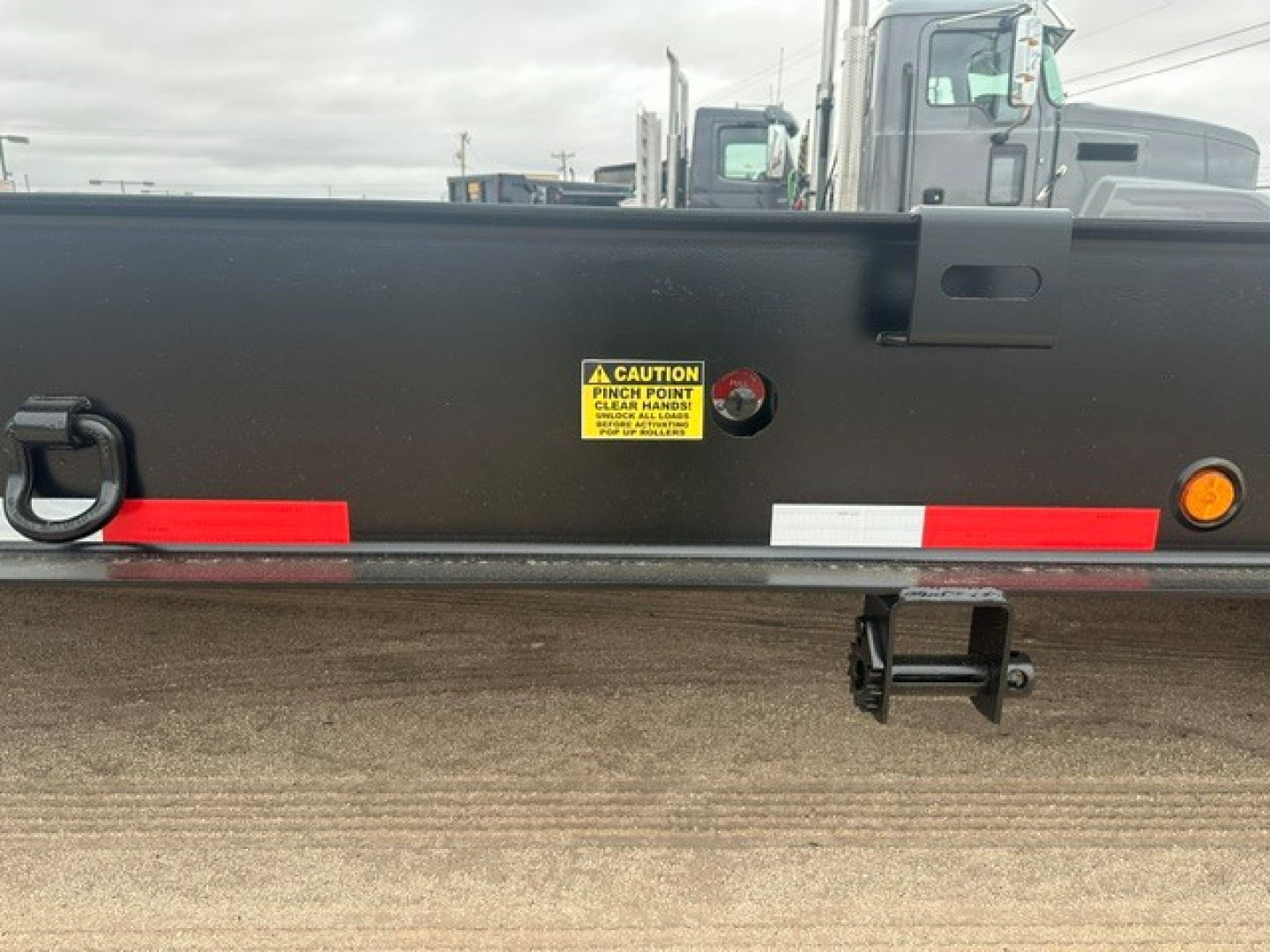 Lowboy trailer rear side with reflective tape, safety sticker, and mounting hardware. Yellow caution label reads CAUTION PINCH POINT ...; black steel surface with red and white reflective strips.