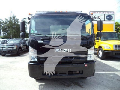 Isuzu truck front view in a dealership lot showing Isuzu emblem on grille and large windshield with side mirrors; surrounding vehicles and dealership sign in background.