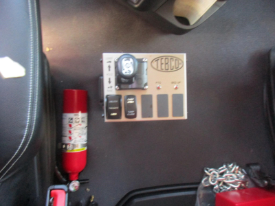 Truck interior panel showing a Terco? control box with switches and a gauge, a red fire extinguisher, and chain on dark surface.