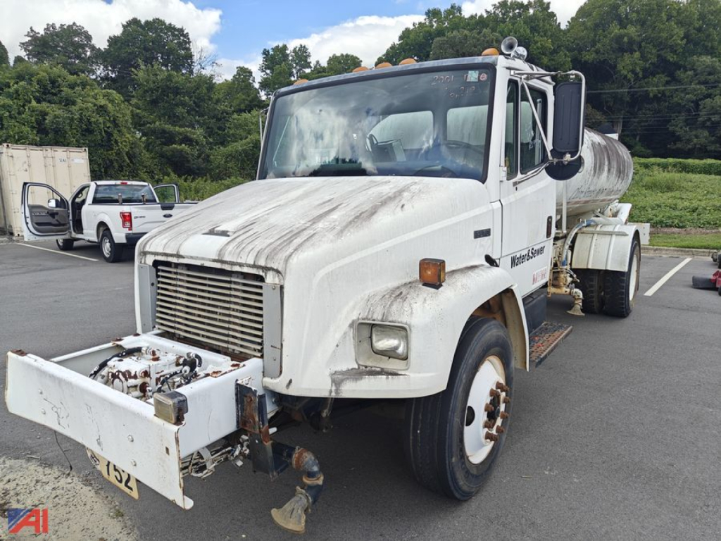 White Freightliner FL70 tanker truck in a parking lot, front-mounted pump and hose assembly visible, with tank behind cab.