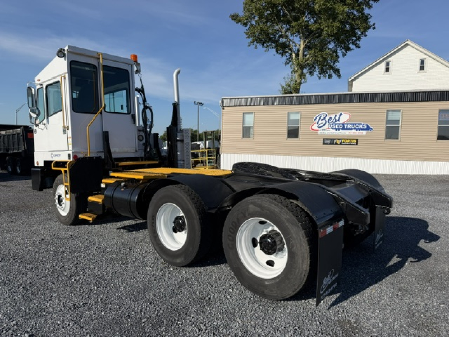 Truck chassis with attached cab and dual rear axles in yard, white cab with black accents, yellow frame, and storage area; sign in background reads Best Used Trucks.